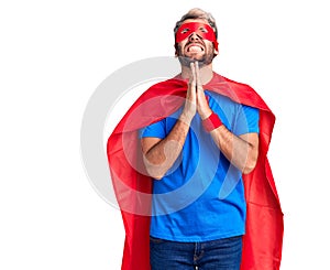 Young blond man wearing super hero custome begging and praying with hands together with hope expression on face very emotional and