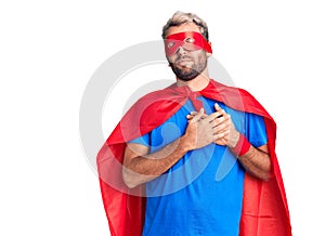 Young blond man wearing super hero custome smiling with hands on chest with closed eyes and grateful gesture on face