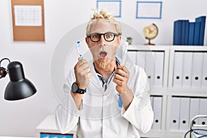 Young blond man working at dentist clinic holding electric toothbrush and teethbrush afraid and shocked with surprise and amazed