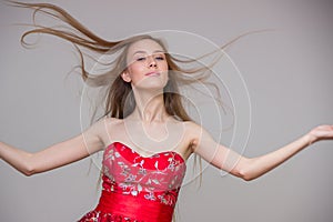 Young blond model with long hair in a smart red dress smiling and having fun on a gray background in the studio