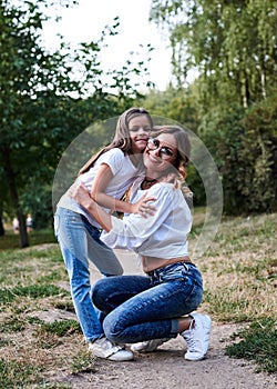 Young blond mother hugging her brunette daughter in the park in summer. Woman and girl, wearing blue jeans and white t-shirts,