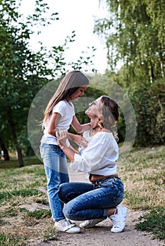 Young blond mother hugging her brunette daughter in the park in summer. Woman and girl, wearing blue jeans and white t-shirts,