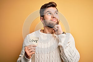 Young blond romantic man with beard and blue eyes holding paper with love message serious face thinking about question, very