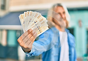 Young blond scandinavian man smiling happy holding danmark 100 kroner banknotes at the city