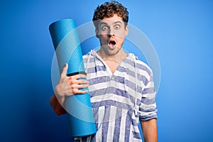 Young blond sporty man with curly hair holding mat to do yoga over isolated blue background scared in shock with a surprise face,