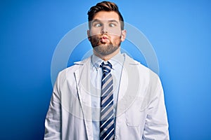 Young blond therapist man with beard and blue eyes wearing coat and tie over background making fish face with lips, crazy and