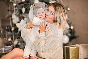 Young blond woman in beige long sweater hugging little baby boy in Christmas suit like a rabbit in white Christmas studio