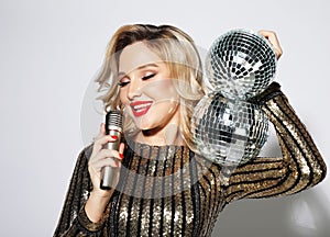Young blond woman dressed in evening dress holding a microphone and disco ball, singing and smiling