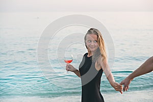 Young blond woman with glass of rose wine holding man's hand on beach by the sea at sunset. Alanya, Turkey.