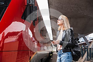 Young blond woman in jeans, shirt and leather jacket wearing bag and sunglass, embarking red modern speed train on train