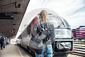 Young blond woman in jeans, shirt and leather jacket wearing bag and sunglass, embarking red modern speed train on train