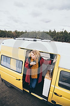 Young blond woman looking out of camper van with solar panel on the roof top and pine forest on the background
