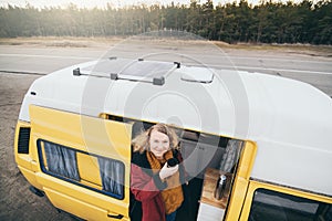 Young blond woman looking out of camper van with solar panel on the roof top and pine forest on the background