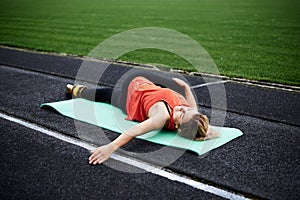 Young blond woman, lying on green yoga mat, doing reclined spinal twist pose. Pretty sportswoman in orange top and black leggings