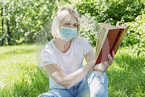 A young blond woman in a medical mask is sitting on the grass with a book in the park. Camping on a sunny day. Precautions during