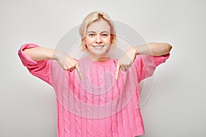 Young blond woman in pink sweater pointing down finger, demonstrating empty space for product or text