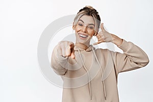Young blond woman pointing finger at camera, showing mobile phone gesture, call me sign, smiling happy, standing over
