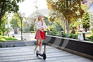 Young blond woman, riding on electric scooter in city center. Full-length portrait of female, wearing red skirt and white top.