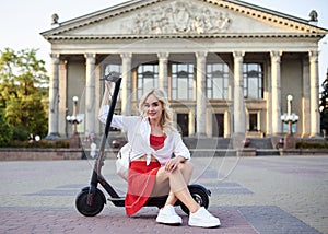 Young blond woman, sitting on black electric scooter in city center in front of old historical building with pillars. Summer