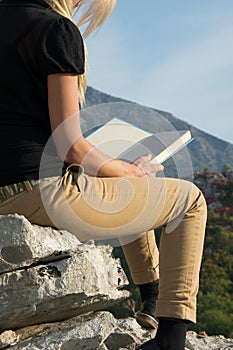 Young blond woman sitting on the edge of the mountain cliff reading a book against beautiful mountains peak