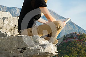 Young blond woman sitting on the edge of the mountain cliff reading a book against beautiful mountains peak