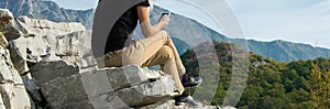 Young blond woman sitting on the edge of the mountain cliff using the smartphone. Technology outdoors lifestyle/travel t