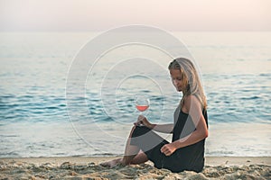 Young blond woman sitting with glass of rose wine on beach by the sea at sunset.