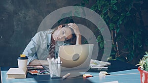 Young blond woman is sitting in office working with computer. She is tired after long day at work