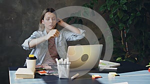 Young blond woman is sitting in office working with computer. She is tired so she is touching her face and hair, rubbing