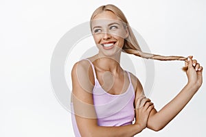Young blond woman smiling and laughing, holding strong healthy hair, standing in casual tank-top over white background
