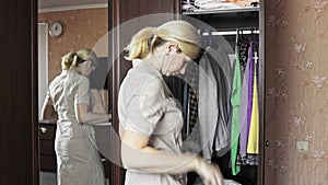 Young blond woman standing in front of an opened wardrobe at home and unbutton her home dress in order to change clothes