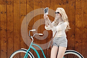 Young blond woman standing near a green vintage bicycle holding photos and smiling, warm, tonning