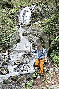 Young blond woman with stick in her hand going down stones along waterfall in the mountain forest