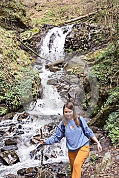 Young blond woman with stick in her hand going down stones along waterfall in the mountain forest