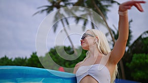 Young blond woman surfer in bikini carrying surfboard walks to ocean and stops looking at waves on tropical beach on