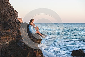 Young blond woman tourist in blue dress sittig on rocks by the sea at sunset. Alanya, Mediterranean region, Turkey.