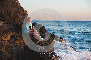 Young blond woman tourist sittig on rocks by the sea at sunset with bottle of lemonade. Alanya, Mediterranean region