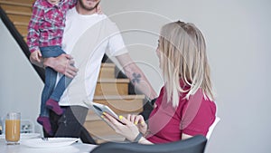 Young blond woman using tablet as her husband coming with daughter on hands. Portrait of happy Caucasian lady spending