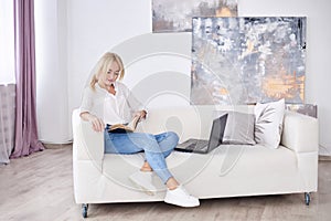 Young blond woman, wearing casual jeans and white top, sitting on sofa, working on design project on laptop. Freelancer employed