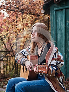 Young blond woman, wearing colorful cardigan, holding acoustic guitar, sitting on a bench in front of old green wooden hut in the