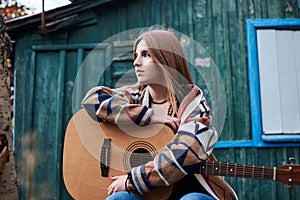 Young blond woman, wearing colorful cardigan, holding acoustic guitar, sitting on a bench in front of old green wooden hut in the