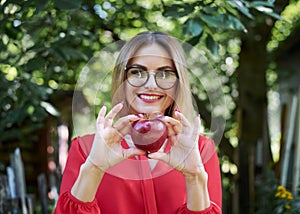 Young blond woman, wearing eyeglasses and red blouse, holding red claret apple in her hand. Close-up picture of female student in