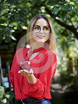 Young blond woman, wearing eyeglasses and red blouse, holding red claret apple in her hand. Close-up picture of female student in