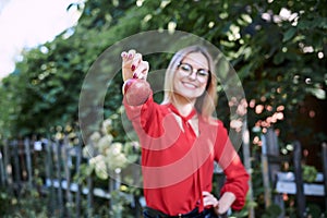 Young blond woman, wearing eyeglasses and red blouse, holding red claret apple in her hand. Close-up picture of female student