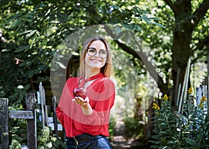 Young blond woman, wearing eyeglasses and red blouse, holding red claret apple in her hand. Three-quarter picture of female