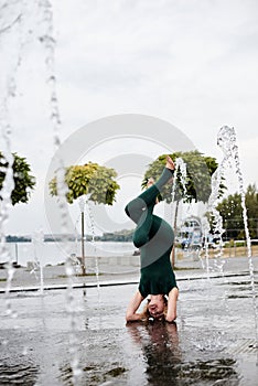 Young blond woman, wearing green overall with bare feet doing yoga pose in fountain near city lake in summer. Yoga practice.