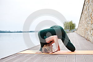 Young blond woman, wearing green overall with bare feet doing yoga pose on orange mat near city lake in summer. Yoga practice.