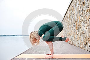 Young blond woman, wearing green overall with bare feet doing yoga pose on orange mat near city lake in summer. Yoga practice.
