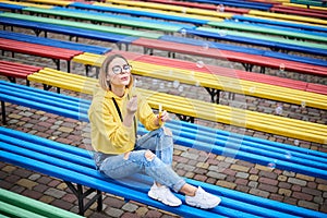 Young blond woman, wearing yellow hoody, blue jeans and eyeglasses, sitting on colorful bench in city urban park in summer.