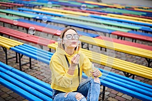 Young blond woman, wearing yellow hoody, blue jeans and eyeglasses, sitting on colorful bench in city urban park in summer.